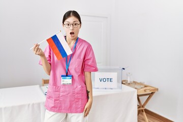 Sticker - Young asian nurse woman at political campaign election holding russia flag scared and amazed with open mouth for surprise, disbelief face