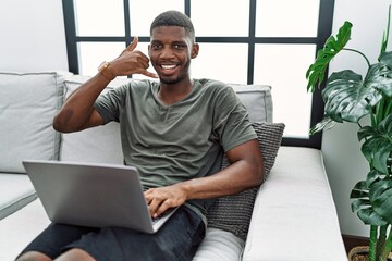 Wall Mural - Young african american man using laptop at home sitting on the sofa smiling doing phone gesture with hand and fingers like talking on the telephone. communicating concepts.