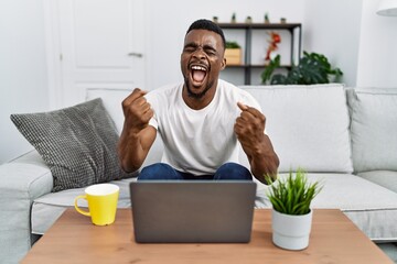 Canvas Print - Young african man using laptop at home very happy and excited doing winner gesture with arms raised, smiling and screaming for success. celebration concept.