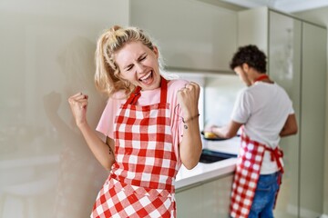 Sticker - Young caucasian woman wearing apron and husband doing housework washing dishes very happy and excited doing winner gesture with arms raised, smiling and screaming for success. celebration concept.