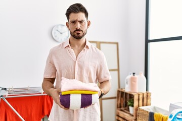 Poster - Young man with beard holding folded laundry after ironing skeptic and nervous, frowning upset because of problem. negative person.