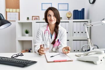 Poster - Young doctor woman holding glass of water and prescription pills smiling looking to the side and staring away thinking.