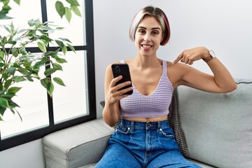 Sticker - Young beautiful woman using smartphone typing message sitting on the sofa looking confident with smile on face, pointing oneself with fingers proud and happy.