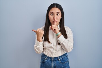 Poster - Young latin woman standing over blue background asking to be quiet with finger on lips pointing with hand to the side. silence and secret concept.