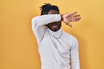 Poster - African man with dreadlocks wearing turtleneck sweater over yellow background covering eyes with arm, looking serious and sad. sightless, hiding and rejection concept