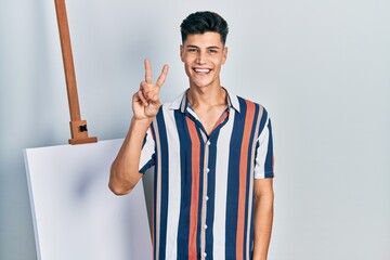 Wall Mural - Young hispanic man standing close to empty canvas showing and pointing up with fingers number two while smiling confident and happy.