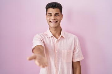 Sticker - Young hispanic man standing over pink background smiling cheerful offering palm hand giving assistance and acceptance.