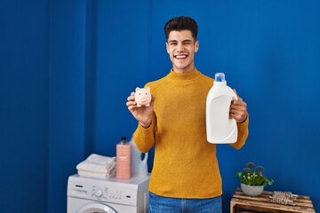 Poster - Young hispanic man holding detergent bottle and piggy bank winking looking at the camera with sexy expression, cheerful and happy face.