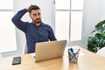 Poster - Young hispanic man with beard working at the office with laptop confuse and wondering about question. uncertain with doubt, thinking with hand on head. pensive concept.