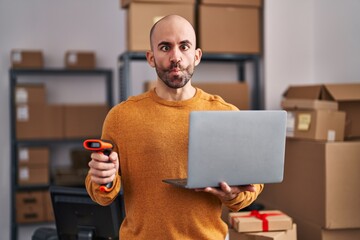 Canvas Print - Young bald man with beard working at small business ecommerce with laptop making fish face with mouth and squinting eyes, crazy and comical.