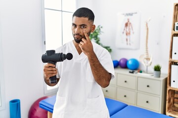 Canvas Print - Young indian physiotherapist holding therapy massage gun at wellness center pointing to the eye watching you gesture, suspicious expression