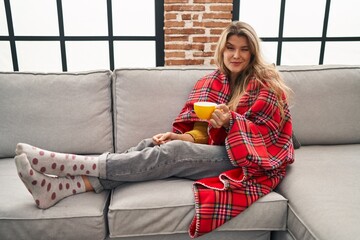 Canvas Print - Young woman sitting on the sofa drinking a coffee at home winking looking at the camera with sexy expression, cheerful and happy face.