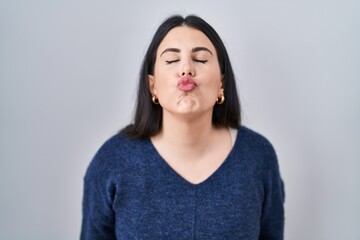 Wall Mural - Young brunette woman standing over isolated background looking at the camera blowing a kiss on air being lovely and sexy. love expression.