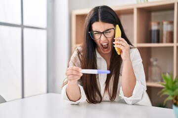 Poster - Young brunette woman holding pregnancy test result speaking on the phone angry and mad screaming frustrated and furious, shouting with anger. rage and aggressive concept.