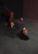 © NASTYA PALEHINA - teen fashion. young brunette woman in a shiny silver bodysuit is lying head down with flirt with hands on chair on the gray wall background in loft interior. fashion concept, free space