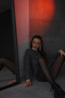 © NASTYA PALEHINA - young girl in shiny silver bodysuit is sitting fashion on floor near mirror with hand on knee on the gray wall background in loft interior and looking down in red light. fashion concept, free space
