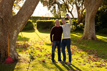 Wall Mural - Image of happy caucasian father and adult son taking selfie in garden