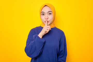 Wall Mural - Serious young Asian Muslim woman dressed in casual sweater showing shh gesture, asks to be quiet isolated over yellow background