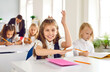 © Studio Romantic - Active schoolgirl. Cute little girl smiling at camera raised her hand while sitting at desk in classroom. Joyful elementary school student sits at desk against background of her classmates.