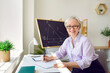 Happy math teacher in the school classroom. Portrait of a beautiful elegant middle aged woman in a shirt and glasses sitting at her working desk, taking some notes, looking at the camera and smiling