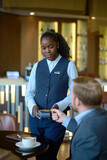 Young African American female worker of hotel with payment terminal standing in front of businessman paying by credit card for coffee