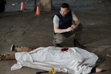 Wall Mural - Young man in bulletproof vest sitting on squats by covered dead body lying on asphalt in parking area during detective investigation