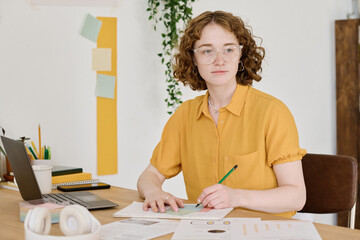 Aufkleber - Young creative woman in eyeglasses and yellow blouse drawing schemes on paper while sitting by workplace and looking forwards