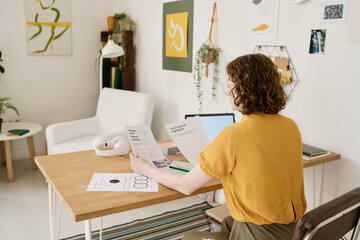 Poster - Back of young female employee in casualwear comparing two financial documents while looking through data by workplace at home