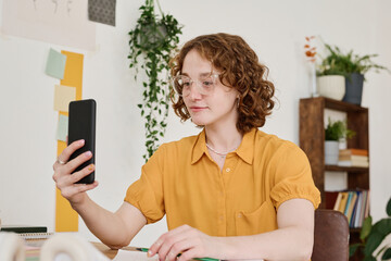 Poster - Young female freelancer in eyeglasses making selfie or communicating in video chat while holding smartphone in front of herself