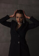 © NASTYA PALEHINA - teen style. young brunette girl in black slim dress-jacket is standing fashion with hands near head and looking straight with flirt on the gray loft wall background. lifestyle concept, free space