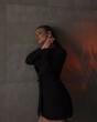 © NASTYA PALEHINA - young girl in black slim dress-jacket  is standing fashion in profile on gray loft wall background with plant in red light and smiling with hands near neck. teen fashion. lifestyle concept, free space