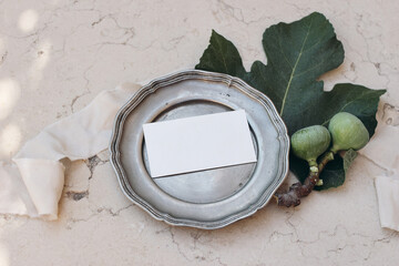 Summer vintage branding, wedding stationery. Mediterranean design. Business card, invitation mock up on old shabby silver plate. Fig fruit, leaves, ribbon. Beige marble background. Flatlay, top view