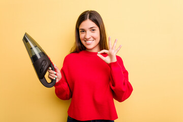 Wall Mural - Young caucasian woman holding a hand vacuum cleaner isolated on yellow background cheerful and confident showing ok gesture.