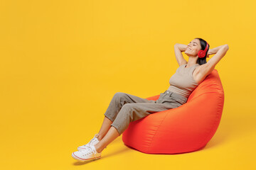 Wall Mural - Full body young happy fun latin woman 30s she wear basic beige tank shirt sit in bag chair headphones listen to calm music hold hands behind neck isolated on plain yellow backround studio portrait