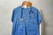 Closeup doctors scrus with stethoscope. Blue surgical smock with stethoscope on wooden hanger on ceramics wall background. Closeup of doctor's scrubs with stethoscope and lab coat on hangers
