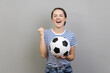 Portrait of woman wearing striped T-shirt screaming holding soccer ball, celebrating victory of favourite football team on championship. Indoor studio shot isolated on gray background.