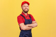 Confident smiling handyman wearing overalls standing with crossed arms, looking at camera, profession of service industry, courier delivery. Indoor studio shot isolated on yellow background.