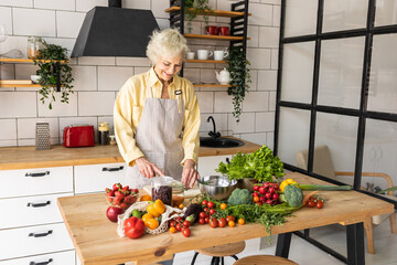 Wall Mural - Attractive senior woman with grey hair cooking healthy food on her kitchen at home. Mature female preparing fresh vegetarian salad with organic ingredients from the market: tomato, cucumber, pepper