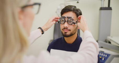 Wall Mural - Optometrist giving male patient an eye test in consultation office. Female ophthalmologist uses spectacle frame with adjustments to check vision of a young Indian man. Optician using trail frame