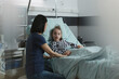 Mother sitting beside hospitalized sick daughter resting in hospital pediatric ward. Caring woman comforting ill little girl under treatment inside children healthcare facility room.
