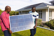 African american senior father and son carrying solar panel while walking in yard against house