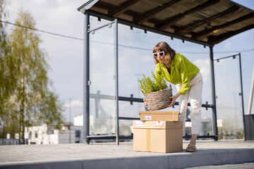 Young woman stands with parcels and flowerpot on a bus stop, waiting for a transportation. Concept of sustainability, delivery and wise use of energy resources