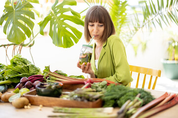 Wall Mural - Young woman drinks from a bottle while sitting by the table full of fresh vegetables, fruits and greens indoors. Concept of healthy vegan food and lifestyle