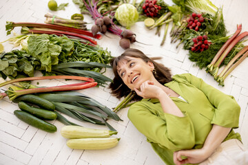 Wall Mural - Portrait of a young cheerful woman with lots of fresh vegetables, fruits and greens above her head, top view. Concept of vegan food and healthy lifestyle