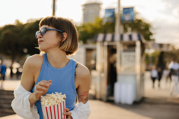 Wall Mural - Satisfied young caucasian brown-haired woman with popcorn walks around city on weekend afternoon. Girl wears sunglasses, blue T-shirt and white shirt. Lifestyle, different emotions, leisure concept.