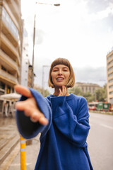 Wall Mural - Beautiful caucasian young woman stretches her hand to camera walking along city street at sunset. Brown-haired with bob haircut wears blue sweatshirt. City life concept