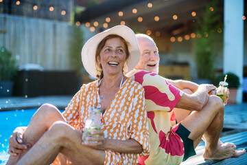 Wall Mural - Happy senior couple enjoying drinks when relaxing and sitting by swimming pool in summer.