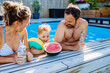 Happy little boy with his parents in backyard swimming pool enjoying refreshments, drinks and watermelon.