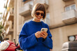 Cheerful caucasian young woman is watching funny video on phone standing on street at daytime. Brown-haired with toothy smile wears sunglasses and sweater. Social media addiction concept