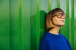 Cute caucasian young lady looking away through sunglasses standing on green wall background. Brown-haired woman with bob haircut wears blue sweatshirt. Rest time concept